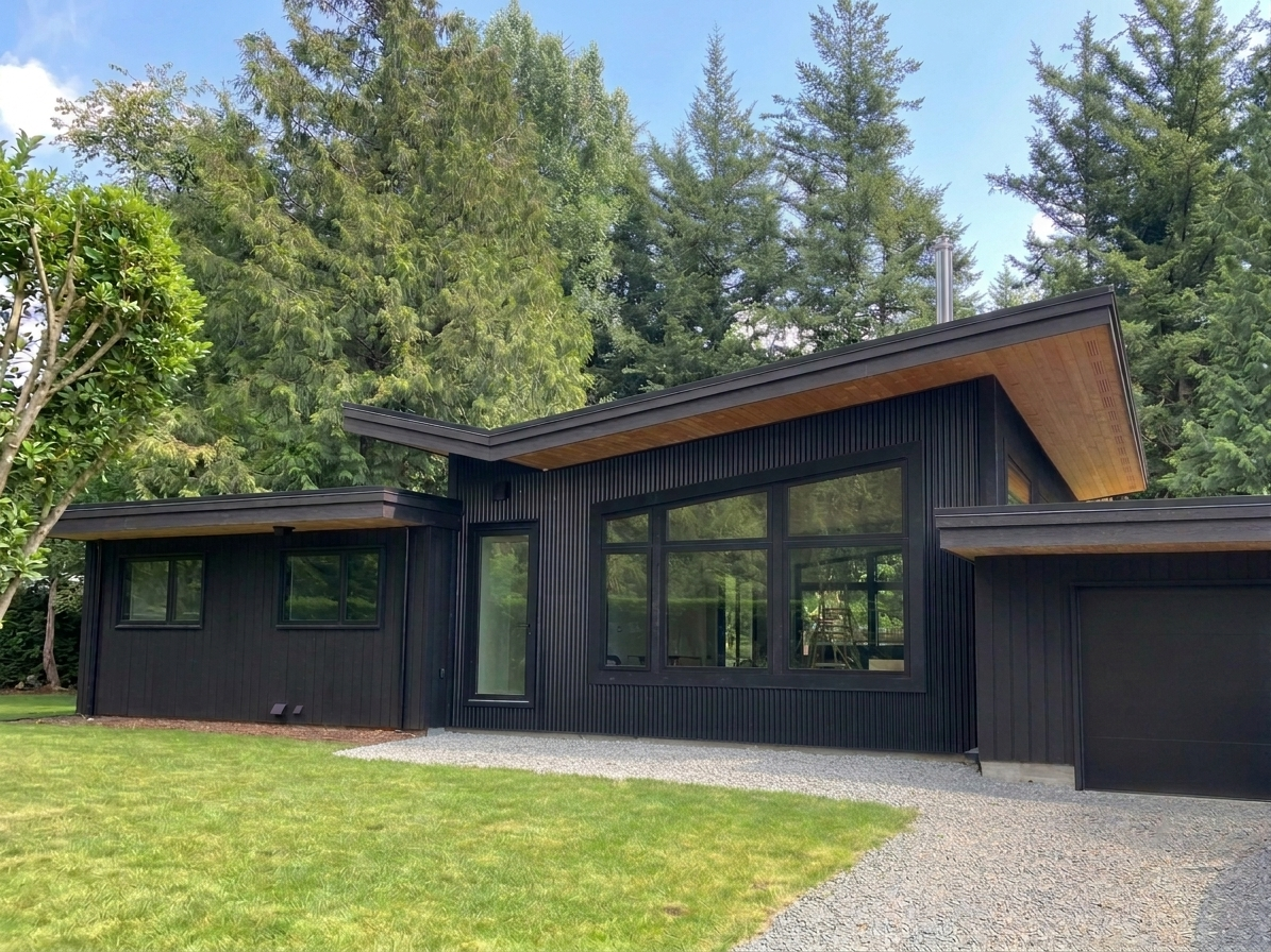 Modern single-story house with dark horizontal siding, large windows, a gravel driveway, green lawn, and tall evergreen trees in the background.